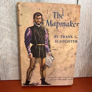 May include: A vintage hardcover book titled The Mapmaker by Frank G. Slaughter. The cover features an illustration of a person in historical attire holding a compass and a scroll against a background of a tan nautical map with grid lines.
