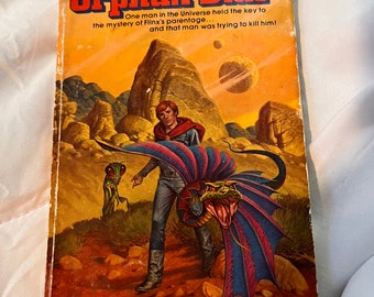May include: A mass market paperback book titled Orphan Star by Alan Dean Foster. The cover features a science fiction illustration of a person in a red cape, a small green insectoid creature, and a large winged serpent on a rocky, orange-hued planet.