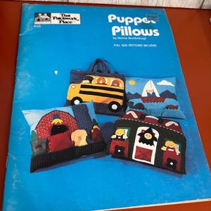 May include: A blue instructional booklet titled Puppet Pillows by Morine Brumbaugh featuring full-size patterns. The cover displays four fabric pillows with appliqued designs including a yellow school bus, a sailboat, a red barn, and a house.