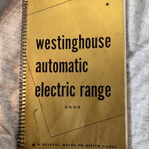 May include: A vintage Westinghouse Automatic Electric Range book with a gold cover and black spiral binding. The cover features the text "Westinghouse automatic electric range" and "A helpful guide to better meals."