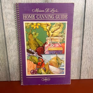 May include: A spiral-bound book titled Miriam B. Loo's HOME CANNING GUIDE with a purple cover. The front features a colorful illustration of fresh produce, including a pear, cherries, and a glass canning jar, alongside a metal ladle.