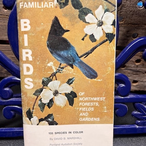 May include: A vintage book titled "Familiar Birds" featuring a blue and black bird perched on a branch with white flowers. The cover is a golden hue with text that reads "Of Northwest Forests, Fields and Gardens" and "133 Species in Color".