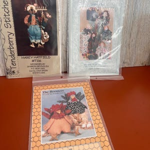 May include: Three craft pattern packets in clear plastic sleeves. One features a rabbit character named Harey Hatfield, another shows multiple doll figures, and the third displays two fabric roosters with the text The Roosters 11 x 14 fun to make roosters.