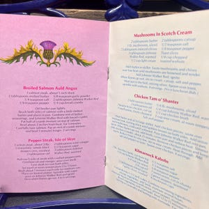 May include: Open cookbook page featuring recipes for Scottish dishes. Recipes include Broiled Salmon Auld Angus, Pepper Steak Isle of Skye, Mushrooms in Scotch Cream, Chicken Tam o' Shanter, and Kilmarnock Kabobs. The page has a thistle illustration.