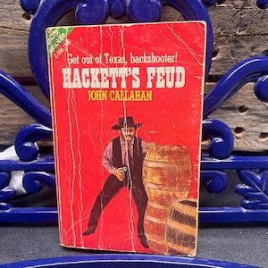 Może przedstawiać: Zabytkowa książka w miękkiej oprawie zatytułowana "Hackett's Feud" autorstwa Johna Callahana. Okładka jest czerwona z ilustracją mężczyzny w kapeluszu kowbojskim i kamizelce, trzymającego broń w pobliżu drewnianych beczek. Tekst "Get out of Texas, backshooter!" znajduje się na górze.