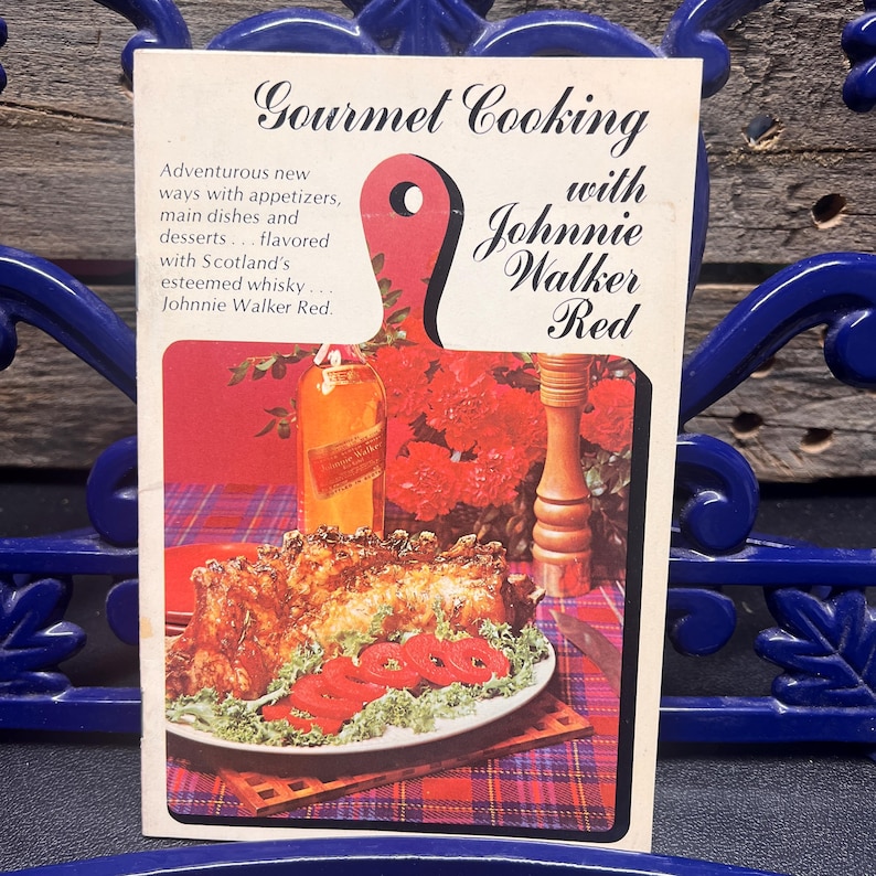 May include: Vintage advertisement for "Gourmet Cooking with Johnnie Walker Red." The image features a red cutting board shape with text and a food illustration. The illustration shows a plate of food, a bottle of Johnnie Walker Red, and red flowers.