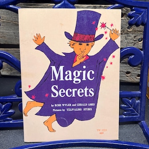 Op de afbeelding: Een vintage boekomslag met de titel "Magic Secrets". De illustratie toont een stripfiguur tovenaar in een paarse cape en hoge hoed, die een toverstaf met een ster vasthoudt. De titel van het boek staat in het wit, met daaronder de namen van de auteurs en de illustrator.