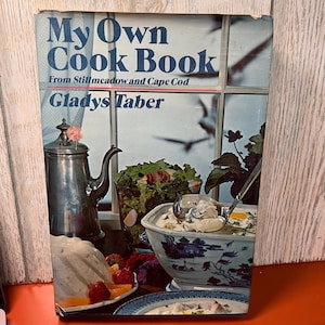 Peut inclure: Un livre de cuisine vintage intitulé "My Own Cook Book" par Gladys Taber. La couverture présente une nature morte avec une cafetière en argent, un verre de salade et un bol de soupe. Le livre est placé devant une fenêtre.