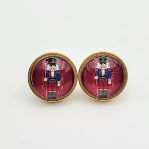May include: A pair of gold-toned earrings with red and white nutcracker images. The nutcracker is wearing a blue and red uniform with a green hat.