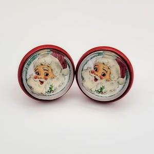 May include: A pair of red and white Christmas earrings featuring a vintage Santa Claus illustration. The earrings are round and have a glossy finish.