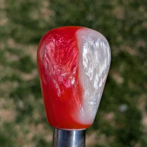 May include: A red and white shift knob with a pearlescent finish. The knob is made of resin and has a smooth, rounded shape.