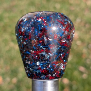 May include: A decorative knob with a clear, glossy finish. The knob is filled with red, white, and blue fragments, creating a patriotic or festive appearance. The knob is mounted on a silver metal post.