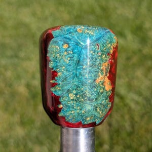 May include: A handcrafted, decorative shift knob with a unique design. The top features a vibrant teal and gold burl wood pattern encased in a glossy, deep red resin. The knob is mounted on a silver metal base, perfect for a car or truck.