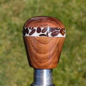 May include: A wooden shift knob with a coffee bean design. The knob is made of dark brown wood with a band of white resin inlaid with brown coffee bean shapes. The knob is mounted on a silver metal shaft.