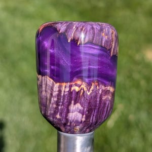 May include: A custom-made, purple and wood-grain shift knob. The knob features a vibrant, shimmering purple resin center, framed by a natural wood grain pattern. The base is a polished silver metal. The shift knob is designed for a car.