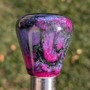 May include: A decorative knob with a glossy finish, featuring a marbled design. The colour palette includes pink, purple, black, and silver. The knob is mounted on a silver metal base. The overall effect is a swirling mix of colours, creating a unique aesthetic.
