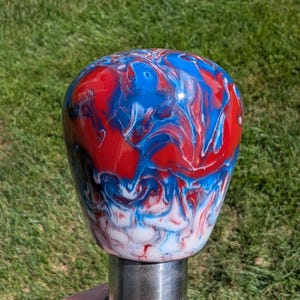 May include: A decorative shift knob with a marbled effect in red, white, and blue. The swirling pattern gives the knob a unique appearance. The knob is mounted on a silver metal base. The overall design is eye-catching.