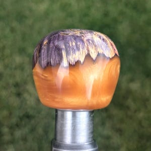 May include: A decorative knob with a metallic silver base and a rounded top. The top is a blend of purple and gold, with a swirling, textured appearance. The knob is likely for a cabinet or drawer.
