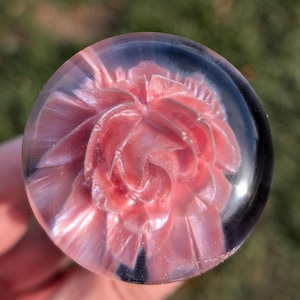 May include: A clear, spherical paperweight featuring a pink, layered floral design. The flower has a detailed, swirling pattern and a pearlescent sheen. The paperweight is held in a hand, with a blurred green background.