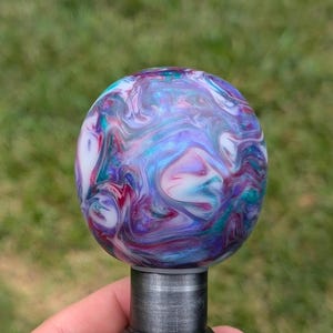 May include: A colorful, swirling, abstract design on a round, white, resin gear shift knob. The knob is mounted on a silver metal shaft.