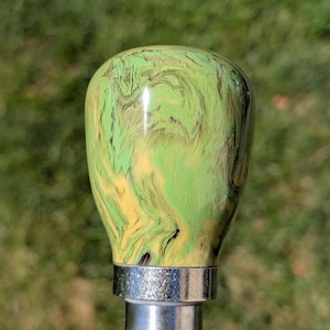 May include: A decorative cane handle with a swirling pattern of green, yellow, and black. The handle is attached to a silver metal base. The background is blurred green.