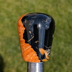 May include: A shift knob with a glossy black and orange design. The top is black, and the side features an orange, wood-grain pattern. It's mounted on a silver metal rod, with a blurred green background. The knob's design is eye-catching.