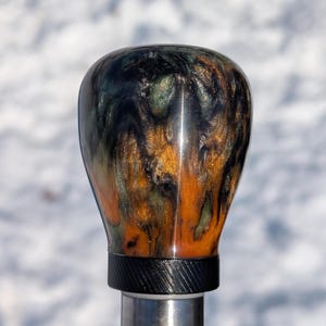 Peut inclure: Un pommeau de levier de vitesses personnalisé avec un design tourbillonnant unique. Le pommeau présente un mélange de résine noire, dorée et verte, créant un effet marbré. La base est noire avec un motif texturé, sur une tige métallique argentée.