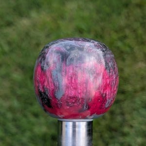 May include: A decorative, spherical object with a swirling pattern of pink, black, and gray colors. The sphere is mounted on a silver metal post. The background is blurred green grass.