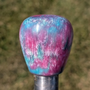 May include: A gear shift knob with a smooth, rounded top featuring a swirling, marbled pattern of vibrant magenta, bright cyan, and pearlescent white. The base of the knob is a cylindrical, metallic silver component with a polished, reflective finish.
