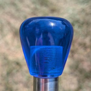 May include: A blue, teardrop-shaped glass object on a silver metal post. The glass is a deep, transparent blue, with a rounded top and a cylindrical base. The background is blurred, suggesting an outdoor setting.