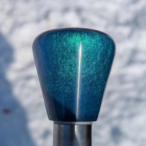 May include: A teal and blue, pearlescent, tapered object with a silver base. The top of the object is rounded. The surface has a glittery, speckled appearance. The background is out of focus, with white and gray tones.