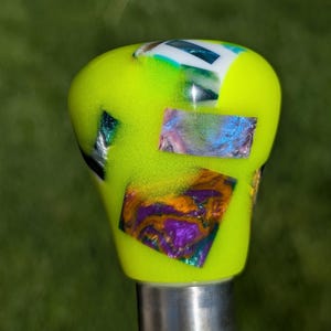 May include: A neon green shift knob with iridescent, rectangular accents in shades of purple, blue, and orange. The knob is mounted on a silver metal shaft, with a blurred green background.