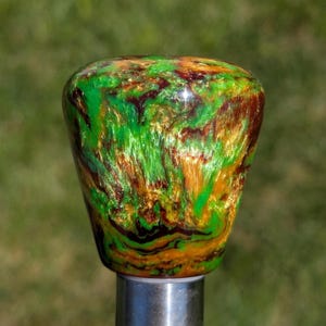 May include: A decorative knob with a vibrant, swirled pattern of green, gold, and brown. The knob is mounted on a silver metal base, set against a blurred green background.
