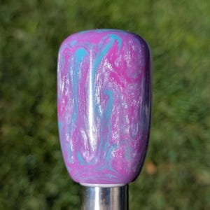 May include: A decorative bottle stopper with a swirling pattern of purple, teal, and white. The stopper has a rounded top and a silver base, perfect for wine bottles or other similar containers.