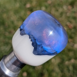 May include: A decorative knob with a galaxy-inspired design. The top section has swirling blue and purple colours with glitter, like a galaxy. The base is white, and the knob is attached to a silver metal stem. A unique, handcrafted item.
