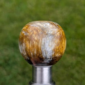 May include: A decorative, spherical knob with a swirling pattern of gold, silver, and brown hues. The knob is mounted on a silver-colored metal base, set against a blurred green background.