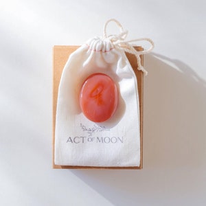 May include: A small, pink, round stone with a symbol on it is in a white drawstring bag. The bag is on top of a brown box with the text "ACT of MOON" printed on it.