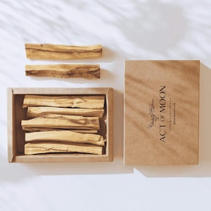 May include: A collection of Palo Santo sticks, some arranged in a rectangular box, others placed separately. A cardboard box with the text "ACT OF MOON" and website address is also present. The sticks are light brown.