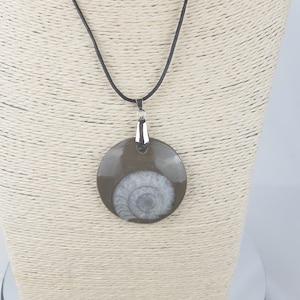 May include: A brown and white ammonite fossil pendant on a black leather cord necklace. The pendant is oval shaped and has a silver bail.