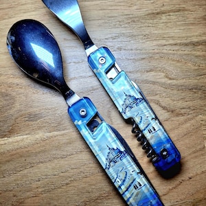 May include: Two-piece camping utensil set with a spoon and a fork. The handles feature a blue and white landscape design. The fork has four tines. The spoon and fork are made of a shiny, dark metal. The handles have a blue button closure.