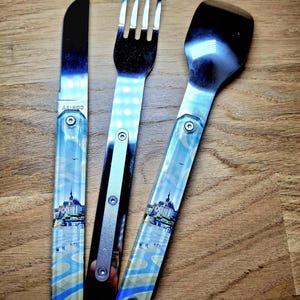 May include: A stainless steel knife, fork, and spatula set. The handles feature a scenic design with a building and people. The knife has the word "AKINOD" printed on it. The utensils are on a wooden surface.