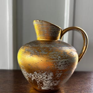 May include: A small, gold-colored pitcher with a rounded body and a handle. The pitcher has a distressed, textured finish with white and silver accents. The spout is slightly angled, and the handle is curved.