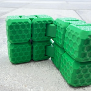 May include: A bright green fidget toy constructed from multiple interconnected cubes. Each cube face features a raised hexagonal honeycomb pattern. The object is articulated, allowing the segments to fold and pivot into various geometric shapes.