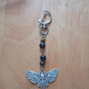 May include: A silver moth-shaped keyring with detailed patterns, suspended from a silver clasp. The keyring includes a string of dark, round beads. The moth design incorporates a skull. The background is a wooden surface.