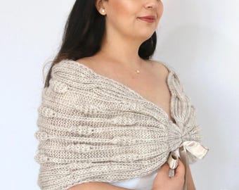 May include: A beige knitted shawl with a textured pattern and a satin ribbon tie. The shawl is worn over a white dress. A white floral bouquet is held in the hand. A decorative headband is worn.