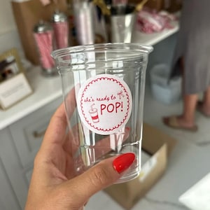 以下が含まれることがあります： 「she's ready to POP!」と書かれた赤と白のステッカーが付いた透明なプラスチックカップ。ステッカーには赤いソーダカップのイラストが描かれています。赤いマニキュアを塗った人がカップを持っています。