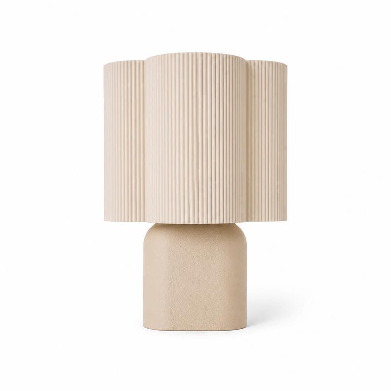May include: A table lamp featuring a beige, vertically ribbed, scalloped cylindrical shade atop a smooth, solid, rounded rectangular base. The lamp has a uniform light tan color throughout, with a distinct contrast between the textured shade and the matte base.