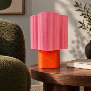 May include: A table lamp featuring a bright pink, vertically ribbed cylindrical shade atop a solid, smooth orange base. The lamp has a distinct two-tone color scheme with a textured upper section and a matte, rounded lower support structure.