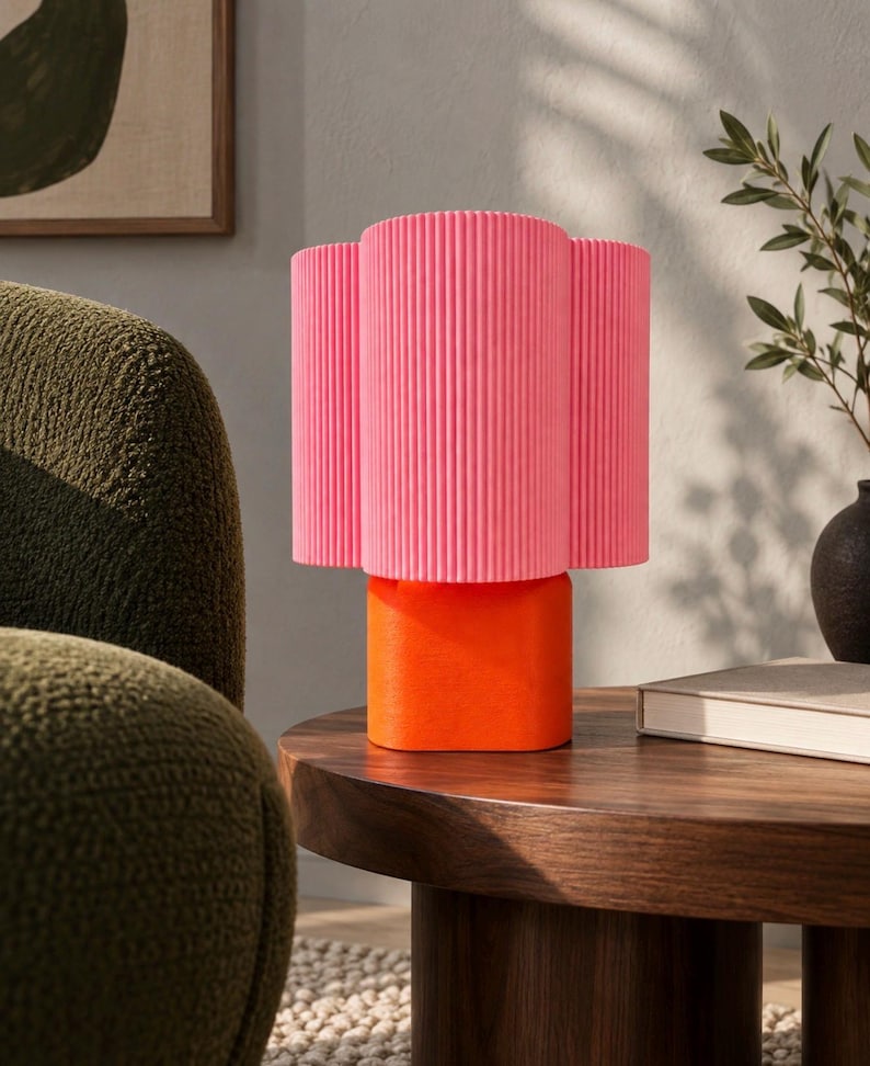 May include: A table lamp featuring a bright pink, vertically ribbed cylindrical shade atop a solid, smooth orange base. The lamp has a distinct two-tone color scheme with a textured upper section and a matte, rounded lower support structure.