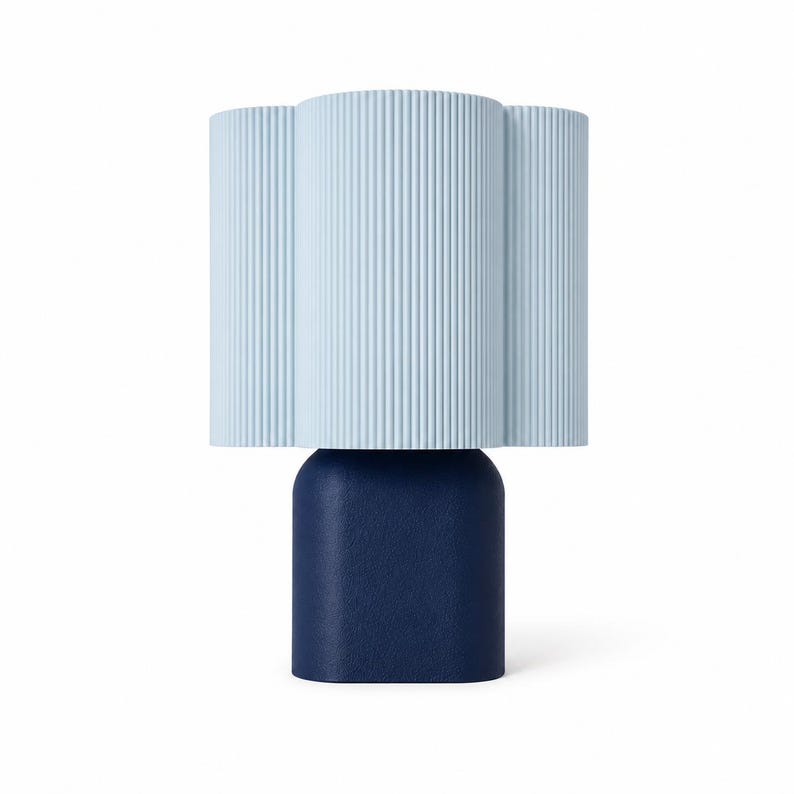 May include: A table lamp featuring a light blue, vertically ribbed, cloud-shaped shade positioned atop a solid, dark navy blue, rounded rectangular base. The lamp has a clean, two-tone design with a textured finish on the base.